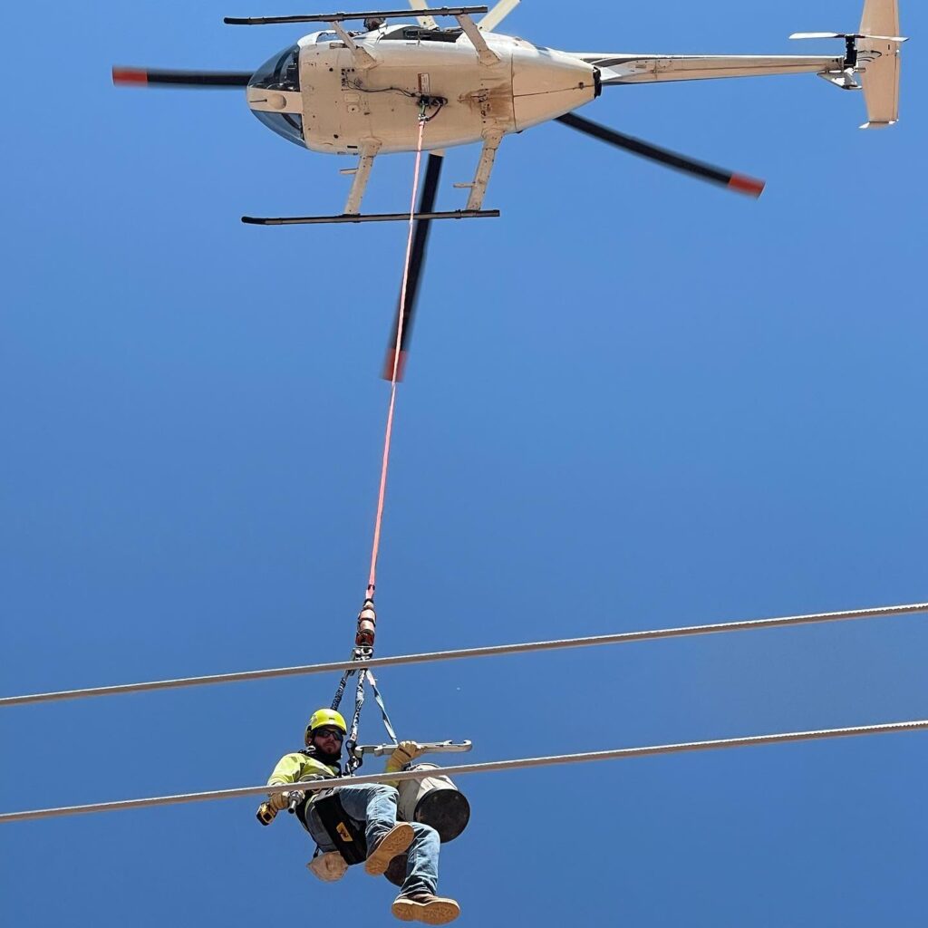 Aerial lineman on a longline beneath a helicopter repairing a power line.