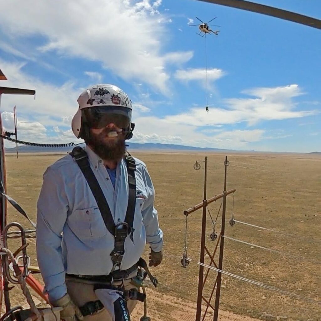 Utility lineman working on a transmission structure while a helicopter performs a longline operation in the background.