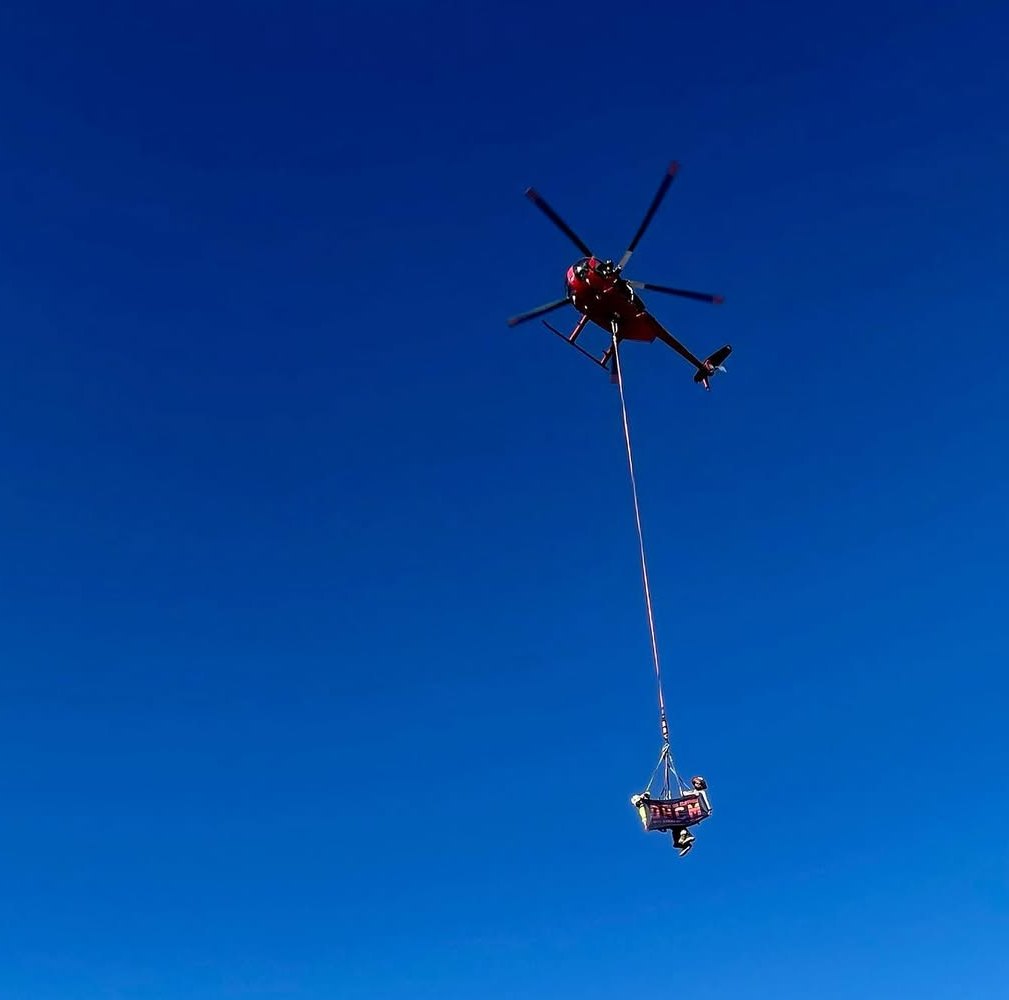 Utility helicopter lifting an aerial platform on a longline against a clear blue sky.