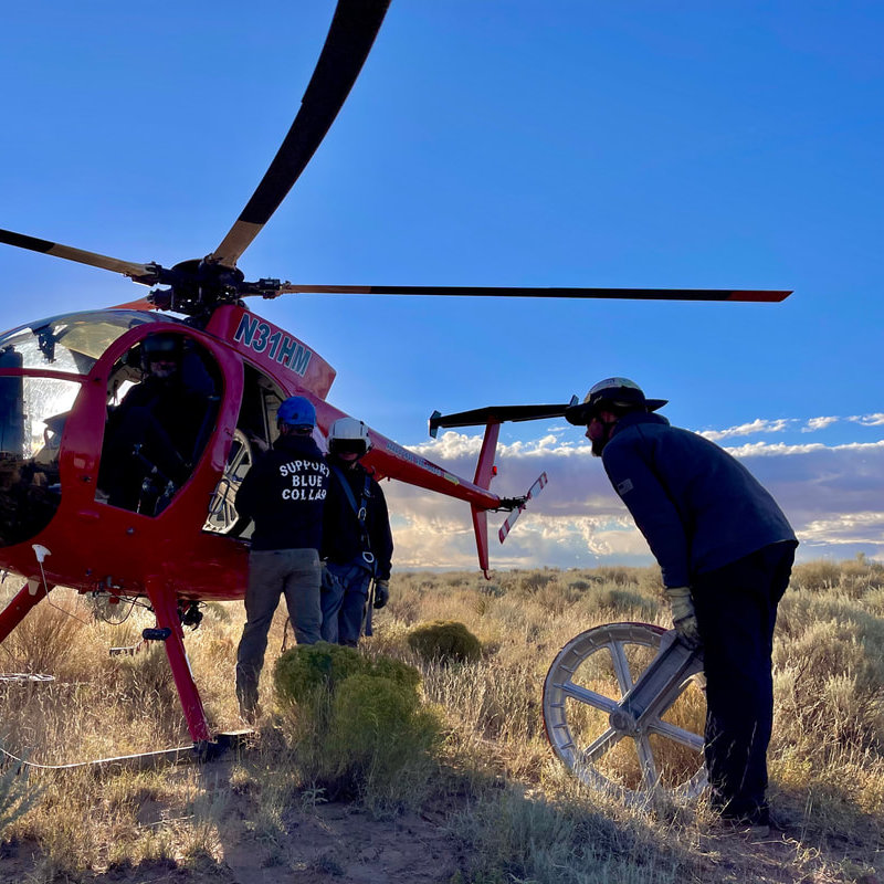 Crew preparing a red utility helicopter for powerline work as a lineman rolls a large conductor pulley across desert terrain.