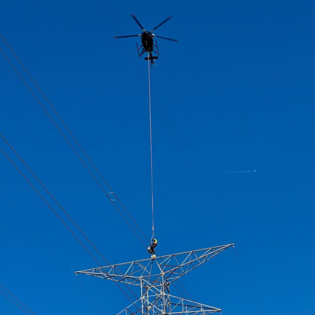 Helicopter performing a Human External Cargo (HEC) lift as a lineman is lowered onto a high-voltage transmission tower.