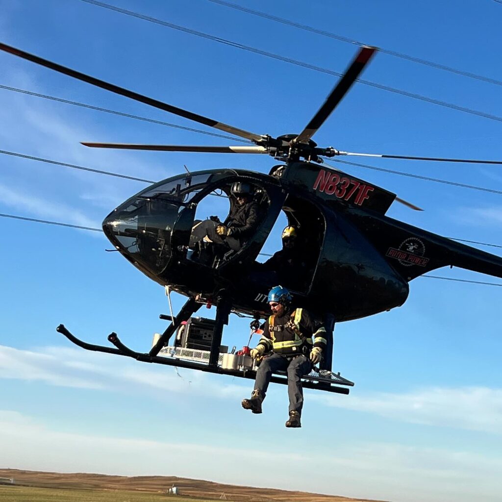 Utility helicopter preparing for HEC transport with a lineman secured on the external platform for a powerline structure repair.